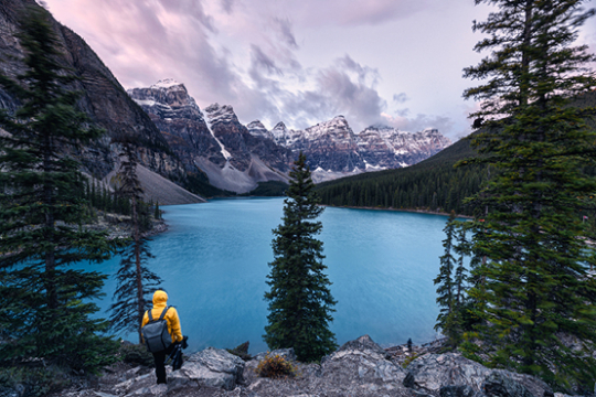 Person in gelber Jacke steht mit dem R&uuml;cken zur Kamera und blickt auf einen gr&uuml;n-blauen See in Kanada umgeben von Bergen